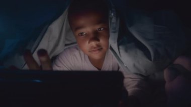 Pijamalı Afrikalı Amerikalı bir çocuğun el feneriyle battaniyenin altında yattığı ve geceleri dijital tablet kullandığı orta boy fotoğraf.