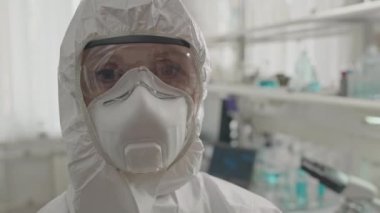 Beyaz, koruyucu tulum giyen, güvenlik gözlüğü takan ve içeride kameraya bakarken solunum maskesi takan kadın bilim adamı portresi.