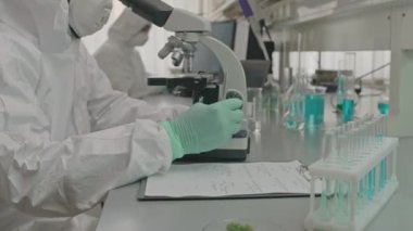 Modern araştırma laboratuvarında meslektaşımla çalışırken mikroskop kullanarak koruyucu üniformalı siyah erkek kimyagerin belini kaldırın.
