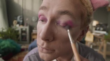 Pembe göz farlarını fırçayla sürerken kameraya bakan beyaz, çekici, eşcinsel bir adamın POV 'u evde gösteri yapmaya hazırlanıyor.