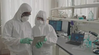 Laboratuvarda dijital tabletle ilgili araştırma verilerine bakarken koruyucu üniformalı ve solunum maskeli iki kimyagerin orta boy görüntüsü.