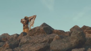 Gün batımında gökyüzünün altında kayanın üstünde uçan süslü şifon eşarbıyla güçlü Afrikalı Amerikalı bir kadının geniş açılı görüntüsü.