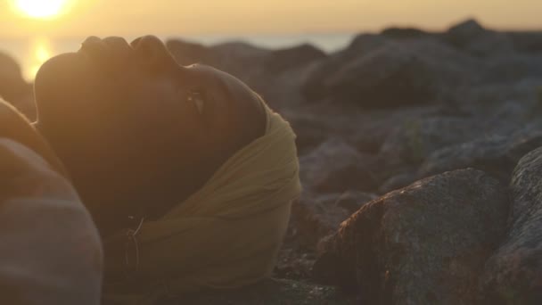 ゴールデンサンセットで一人でビーチストーンに横たわる謎の若いアフリカ系アメリカ人女性のミディアムクローズアップとまっすぐ前を見る — ストック動画