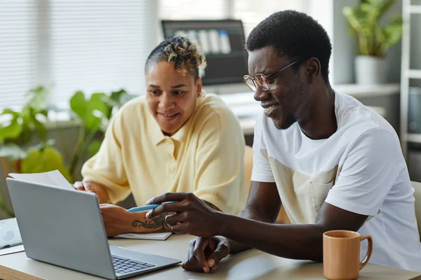 Side view portrait of two smiling black people using laptop together in IT office and wearing casual clothes pointing at screen