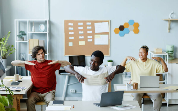 Diverse group of smiling young people stretching muscles as fun warm up at office workplace