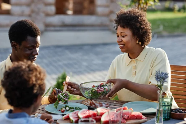 Side view portrait of smiling African American woman serving food to family while enjoying dinner together outdoors