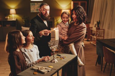 Hanuka kutlamaları sırasında sıcak ev ortamında gümüş menorah mum yakan modern Yahudi aile portresi.