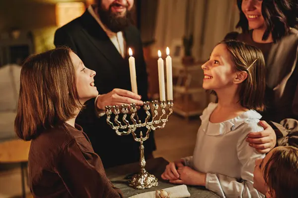Portrait of orthodox jewish family lighting menorah candle together during Hanukkah celebration with focus on two girls smiling