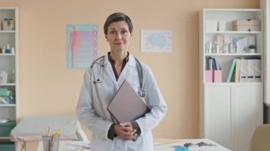 Beyaz laboratuvar önlüğü ve steteskop giymiş orta yaşlı beyaz kadın doktorun modern hastane ofisinde dizüstü bilgisayarla fotoğraf çekerken orta boy portresi.