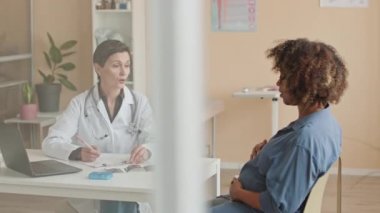 Genç Afrikalı hamile kadın ve beyaz kadın jinekoloğun gündüz vakti klinikte konuştuğu cam bölme duvarı vasıtasıyla.