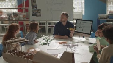 Orta yaşlı bir grup ilkokul çocuğu ve Kafkas erkek öğretmenin çalışma masasında oturup konuştuğu ve uçak modelleri yaptığı bir fotoğraf.