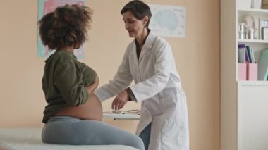 Orta boy beyaz kadın jinekolog ölçü bandı kullanırken klinikteki kanepede oturan genç siyahi hamile kadının göbek ölçüsünü alırken.