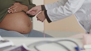 Modern bir klinikte, karnının muayene edilip kadın doktor tarafından ölçüldüğü, tanınamayan hamile bir kadının görüntüsü.