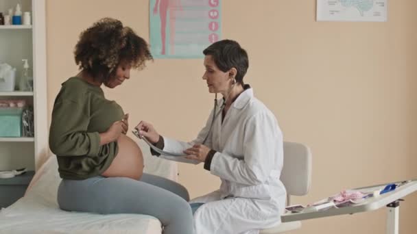 コカシア人女性医師のメディアショット 医療オフィスでステススコープを有する若いアフリカ系アメリカ人女性の妊娠中の胃を聞く — ストック動画