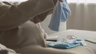 Bebek ultrasonuna bakıp bebek kıyafetlerini katlarken karnını okşayan tanınmayan hamile bir kadının görüntüsü.