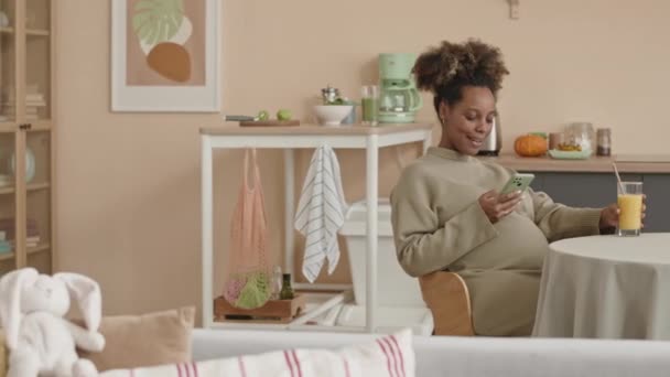 在舒适的公寓里 坐在厨房桌子边喝着新鲜橙汁 用智能手机浏览的年轻漂亮的黑人孕妇的潘中景照片 — 图库视频影像
