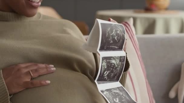 年轻的非洲裔美国孕妇兴奋地抚摸着她的腹部 一边看着婴儿超声波扫描 一边充满爱意和柔情 坐在舒适的家庭氛围中的沙发上 — 图库视频影像
