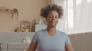 Geniş bir stüdyo dairede yerde yoga meditasyonu yapan rahat siyahi bir kadın.