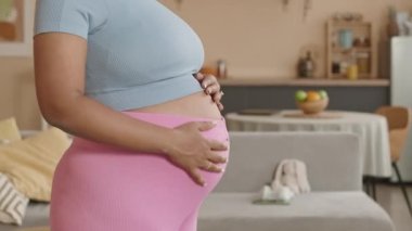 Sıkı pembe taytlar giyen, belini okşayan, evde yoga yapan ve boş vakitlerinde oturma odasında yoga yapan genç ve hamile bir kadını yavaşça yukarı kaldır.