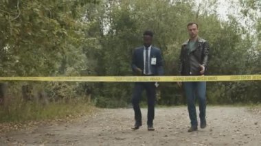 Çoklu etnik çeşitlilikte iki dedektifin barikatı aşıp parkta, siyah plastik torba altında cesede yaklaşırken sohbet etmelerinin tam görüntüsü.
