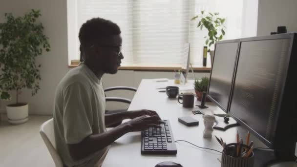 サイドビューの若いアフリカ系アメリカ人の男性ギークは ミニマリストのオープンスペースオフィスで働いている間 2つのモニターの前に座ってプログラムコードを入力する基本的なTシャツと眼鏡を身に着けています — ストック動画