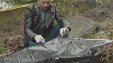 Deri ceket ve eldiven giyen beyaz erkek dedektifin, içinde ceset olan siyah plastik torbayı açarken ve içinde boşaltılmış akıllı telefonu bulunca orta boy fotoğrafı.