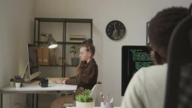 Rastalı kollarını ve sırtını uzatan ve meslektaşıyla açık uzay ofisinde çalışırken masasında kafa rotasyonu yapan yorgun, beyaz kadın programcı.