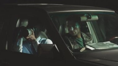 İki etnik çeşitliliğe sahip erkek polisin gece vakti arabayla suçluları takip etmesi ve takip etmesi. Profesyonel kamerada fotoğraf çeken siyah bir erkek polis memuru sürücü koltuğundaki meslektaşlarına gösteriyor.