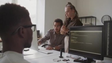 Bilgisayar sunumu ve önplanda siyahi erkek meslektaşlarının program kodlarını yazarken çok ırklı iki meslektaşın tartıştığı orta boy bir fotoğraf.