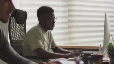 Modern açık uzay ofisindeki bilgisayar monitörlerinin önünde oturan iki çok ırklı erkek yazılım geliştiricisinin belden yukarı görüntüsü