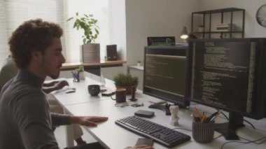 Çift taraflı kıvırcık saçlı yakışıklı iki bilgisayar monitörünün önünde oturuyor ve klavyeye program kodları yazmaya başlıyor.