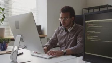 Masaüstü bilgisayarında yazılım geliştirirken kablosuz klavyede yazan konsantre çift ırklı erkek programcı, modern minimalist ofiste çalışıyor.