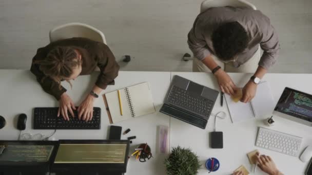 2人のプロのソフトウェア開発者のトップビューは 粘着性のあるノートを見て ホワイトオフィスデスクでデスクトップコンピュータで一緒に座って議論しています — ストック動画