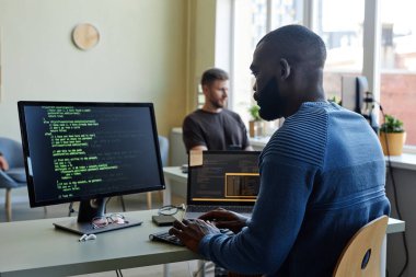 Ofisteki işyerinde çoklu aygıtlarla kod yazan siyah yazılım mühendisinin yan görünüm portresi