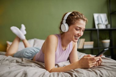 Kablosuz kulaklıklı genç bir kadın yatağında dinleniyor ve akıllı telefonunu kullanarak internette konuşuyor.