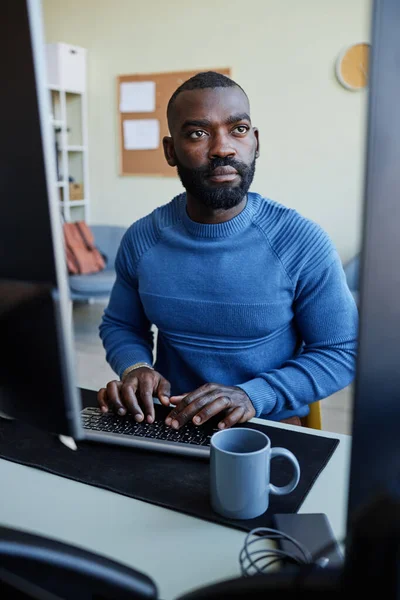 Vertical portrait of focused black man programming software at workplace with multiple computer screens