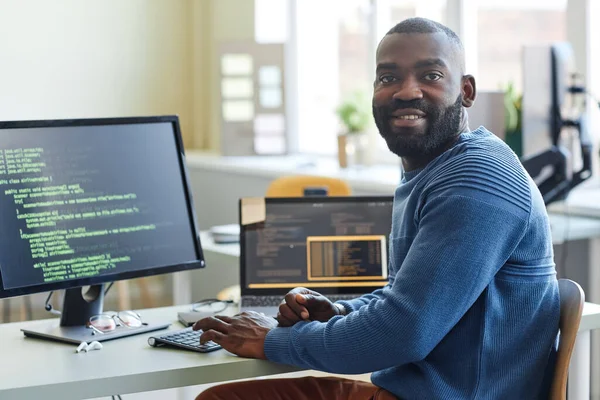 Portrait of black man as computer programmer looking at camera while writing code at office workplace