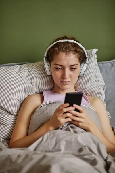 Young girl in wireless headphones listening to audio book on her smartphone while lying on bed in bedroom