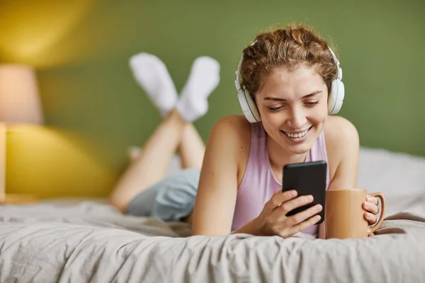 Smiling girl in wireless headphones communicating online using her smartphone and drinking coffee in bed in the morning