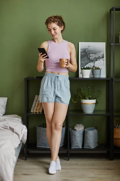 stock image Young woman in domestic clothes communicating online on her smartphone and drinking coffee standing in her bedroom