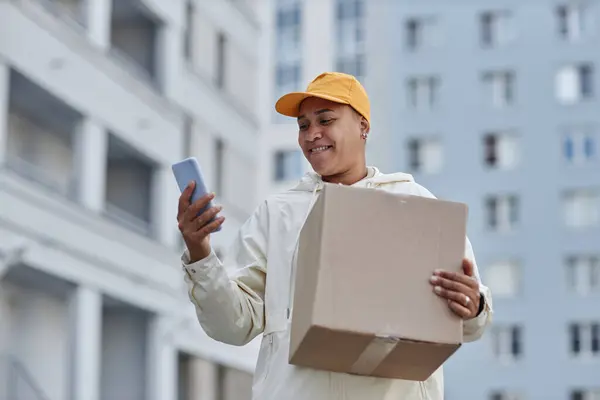Waist up portrait of multiethnic young woman delivering packages in city and using smartphone
