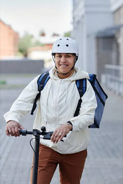 都市の電動スクーターに乗っている間ヘルメットを身に着けている笑顔の食品配達労働者の縦の肖像 — ストック写真