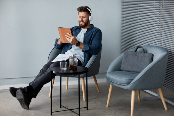 Happy young man in headphones and casualwear using digital tablet while sitting in armchair and communicating in video chat