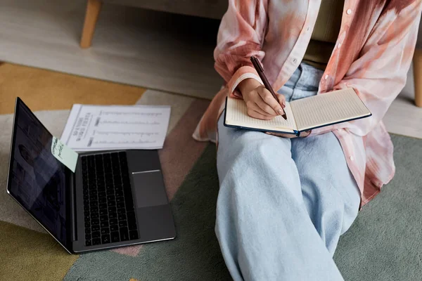 Stock image Close-up of young female student in casualwear sitting on the floor and making notes in notebook during online lesson with tutor