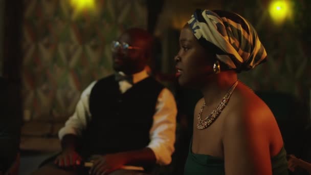 年轻的非洲裔美国女歌手头戴花纹头巾 戴着五彩缤纷的珠项链 与室内爵士乐俱乐部的乐队成员讨论音乐会节目 灯光暗淡 — 图库视频影像