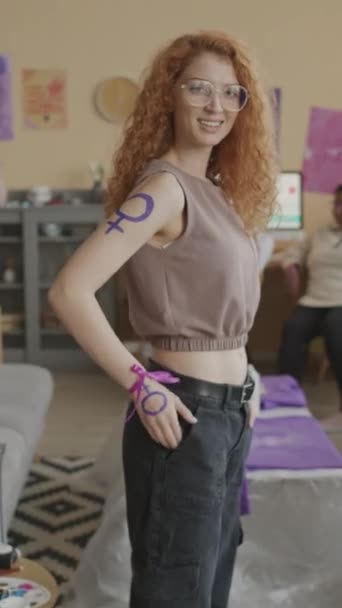 紫のフェミニストのシンボルとリスト上のリボンのカメラのための長いカーリージンジャーの髪を持つ若い白人女性の垂直中型の肖像画は フェミニストのイデオロギーのサポートを示しています — ストック動画