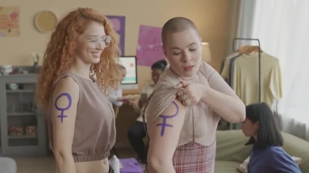 2人の若い白人女性活動家の中央肖像画は 自宅で他の女性とコミュニティーミーティングを行っている間 肩に紫のフェミニストのシンボルを示しています — ストック動画