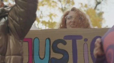 Feminist hareketi ve ideolojiyi destekleyen diğer aktivistlerle açık hava protestoları sırasında elinde karton Adalet Şimdi afişi tutan genç ve aktif feminist kadının orta boy fotoğrafı.