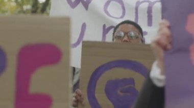 Sokaklarda gerçekleşen protesto toplantısında feminist afişleri ve posterleri olan genç ve aktif kadınların orta boy taçlarını çekip yumruklarını kaldırarak bağırıyorlar.