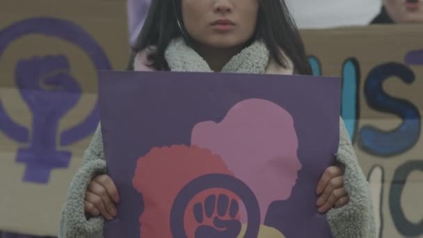 バックグラウンドで叫ぶ多様な女性との屋外デモプロセス中にストリートでカメラのためにポーズ紫のフェミニストのポスターを持つ若いイニシアチブの若いアジア女性のスローモの肖像画を傾ける — ストック動画